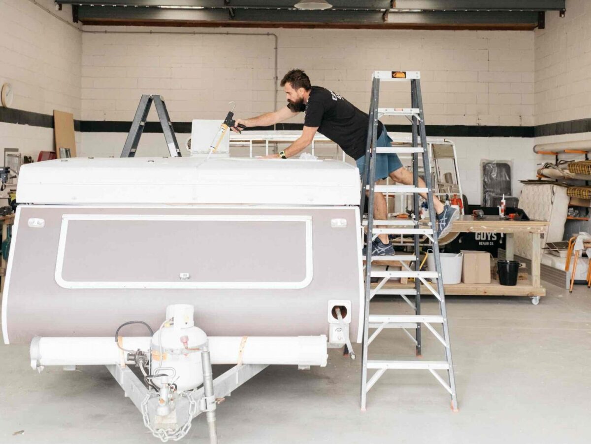 Worker conducting a caravan service on a ladder next to a folded down caravan in a workshop. He is doing a caravan roof reseal.