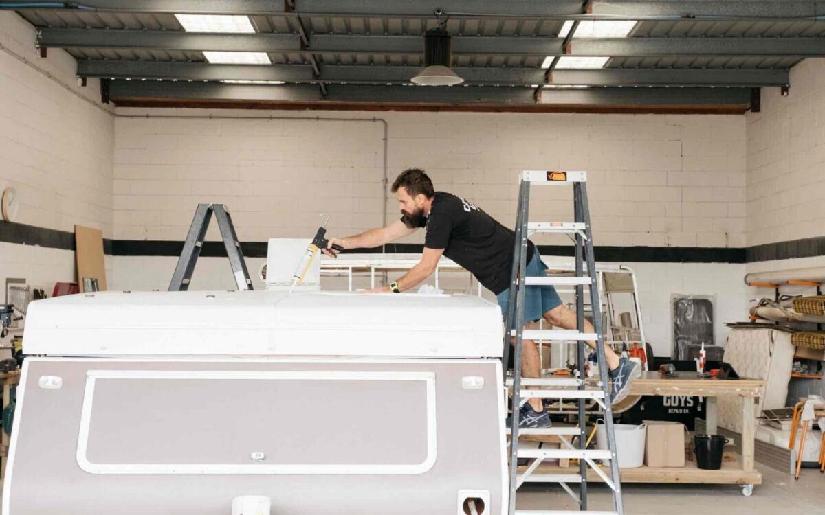 Worker conducting a caravan service on a ladder next to a folded down caravan in a workshop. He is doing a caravan roof reseal.