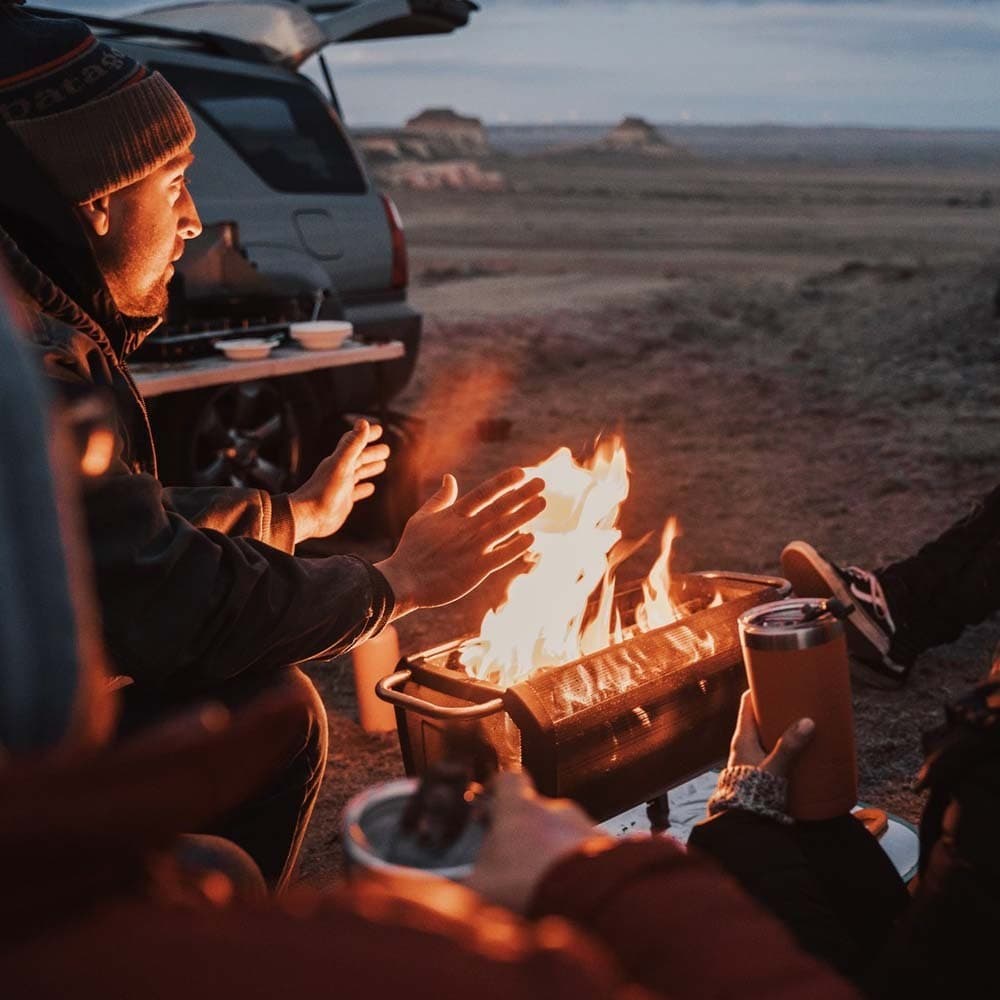Group of campers around a portable fire pit, warming hands and drinking near a vehicle at dusk.