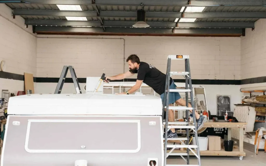 Worker conducting a caravan service on a ladder next to a folded down caravan in a workshop.